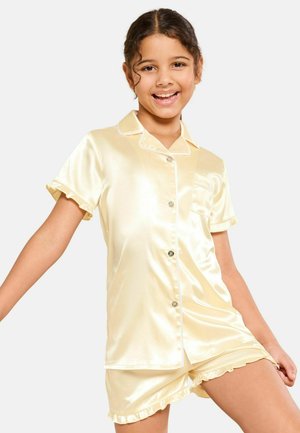 Mosolygós lány sötét göndör hajjal krémszínű szatén rövid ujjú, gombolós ingben és hozzá illő fodros rövidnadrágban fehér háttér előtt.