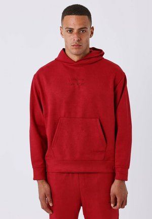 Sweat-shirt à capuche rouge en tissu doux, avec une poche avant et un logo en relief sur la poitrine. Texture lisse, coupe décontractée.