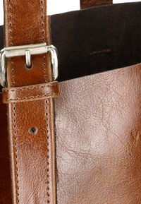 Brązowa skórzana torba tote z gładką teksturą, regulowanym paskiem z metalowymi detalami i przeszytym wykończeniem. Wnętrze wydaje się ciemne.