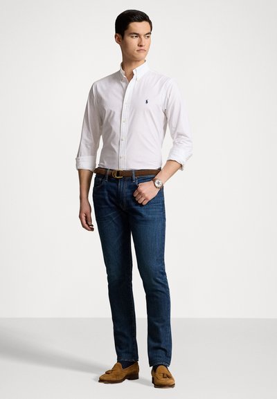 Witte blouse met knopen, blauwe spijkerbroek, een bruine leren riem, tan kleurige moccasinschoenen en een horloge; staande pose met één hand op de heup.