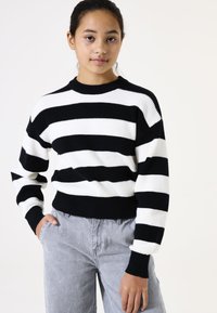 Gestreifter Strickpullover in Schwarz und Weiß mit Rundhalsausschnitt und geripptem Stoff. Kombiniert mit grauen Jeanshosen mit Seitentaschen.