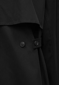 Gros plan sur un tissu noir avec deux boutons noirs et un détail de sangle, montrant la couture et la conception superposée du vêtement.