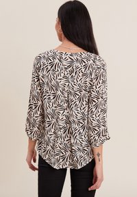 Blusa bianca con motivo a foglia nera e arancione, vestibilità rilassata, maniche 3/4, scollatura a barca, tessuto leggero. Indossata con pantaloni neri.