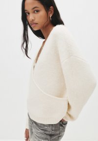Kremowy wełniany sweter z głębokim dekoltem w kształcie litery V, o luźnym kroju i teksturowanym splocie. Połączony z szarymi dżinsami, podkreślającymi ekologiczny materiał.
