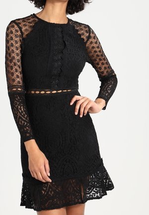 Schwarzes Spitzenkleid mit langen transparenten Ärmeln, Ausschnitt am Bauch und gewelltem Saum über blickdichtem Futter, getragen von einer Person mit Hand in der Hüfte.