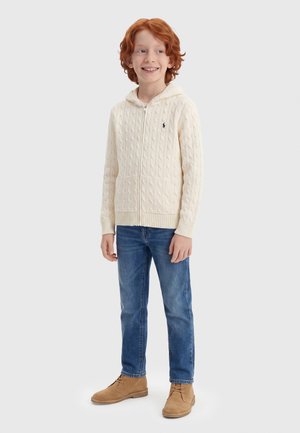 Χαμογελαστό αγόρι με κόκκινα μαλλιά που φοράει κρεμ ζακέτα με φερμουάρ και πλέξη καλωδίων, μπλε τζιν και ταμπά δερμάτινα μπότες με κορδόνια, στέκεται σε λευκό φόντο.