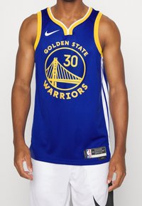 Uomo che indossa una maglia da basket blu dei Golden State Warriors con il numero 30, pantaloni bianchi, in piedi su uno sfondo neutro.