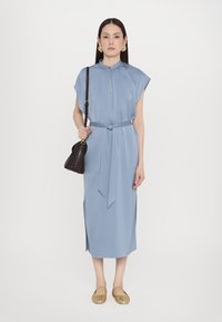 Ανοιχτό μπλε, φόρεμα έως το γόνατο με κοντά μανίκια, ζώνη που δένει στη μέση, πλαινές σχισμές και λεπτομέρεια με κλειδαρότρυπα στον λαιμό. Τα αξεσουάρ περιλαμβάνουν μια μαύρη τσάντα.