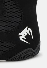 Schwarzer Laufschuh aus einer Kombination von glatten und mesh Materialien, mit einem auffälligen weißen Logo und einer strukturierten Gummisohle.