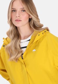 Sárga vízálló dzseki cipzárral és húzózsinóros kapucnival. Fehér esernyő logóval, hálós béléssel és laza szabással.