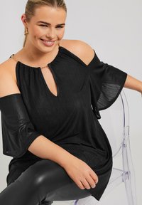 Top nero senza spalle con scollatura a chiave, tessuto texturizzato, maniche svasate e dettagli in metallo dorato. Indossato con leggings neri.