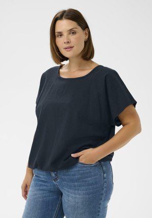 Femeie cu păr castaniu până la umeri, purtând un top lejer bleu închis și blugi albaștri, pozând cu o mână în buzunar pe un fundal alb.