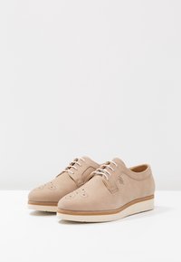Chaussures en cuir beige avec une texture lisse, bout arrondi, perforations à l'avant et lacets blancs. Elles présentent une semelle légère et texturée.