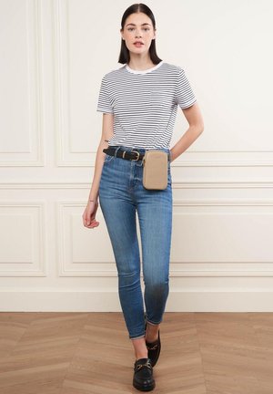 Gestreiftes weißes und dunkelblaues T-Shirt, eng anliegende blaue Jeans, schwarzer Gürtel und schwarze Slipper. Braune Tasche am Gürtel befestigt. Heller Holzboden.