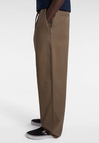 Brown cotton trousers with a relaxed fit, featuring side pockets and an elastic waistband with drawstring. Paired with black shoes.