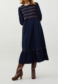 Navyblaues langes Kleid mit Puffärmeln, dekorativer orangefarbener Naht, gerafftem Taillenbund und schwingendem Saum. Braune Absatzstiefel.