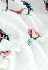 Biely textil s textúrovanými volánmi obsahuje farebnú kvetinovú výšivku v ružovej, červenej a modrej farbe. Hladký materiál kontrastuje s vystúpenými vzormi.