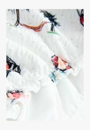 Bílá látka s texturovanými volány má barevné květinové výšivky v růžové, červené a modré. Hladký materiál kontrastuje s vyvýšenými vzory.