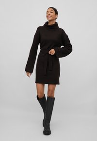 Vestido de malha castanho com colarinho alto, mangas longas e um cinto atado na cintura. Combinado com botas pretas até ao joelho para contraste.