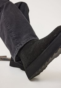 Schwarze Wildleder-Stiefeletten mit kleinen funkelnden Nieten, getragen mit dunkelgrauen Jeans, die die strukturierte Sohle und das Seitenprofil zur Schau stellen.