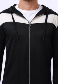 Haut à capuche zippé noir avec une bande blanche sur la poitrine. Il dispose d'une fermeture éclair à l'avant et d'une capuche avec cordon de serrage ajustable. Texture douce, design décontracté.