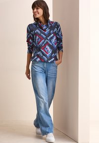 Maglione a fantasia multicolore in blu, rosso e grigio; presenta un collo a coda di rondine. Abbinato a jeans a gamba larga di un blu chiaro e sneaker bianche.