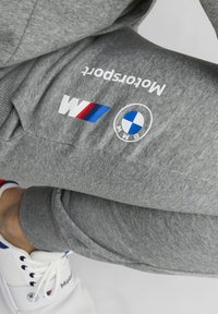 Grå sweatpants med en BMW motorsport-logotyp, blå och röda accentremsor och ribbade muddar, bärs med vita skor.