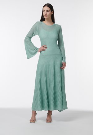 Femme aux cheveux longs et lisses portant une robe en maille côtelée vert clair avec des manches évasées et une jupe plissée, debout avec une main sur la hanche.