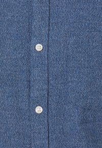 Een button-up overhemd van textuurblauwe stof, met twee witte knopen aan de voorkant en een subtiel geweven patroon.