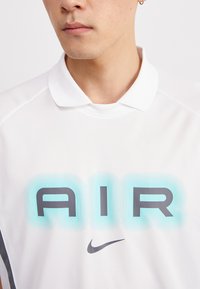 Biele polo tričko so štruktúrovaným golierom, s veľkým potlačou "AIR" v modrej a sivej farbe. Logo Nike je diskrétne zobrazené pod textom.