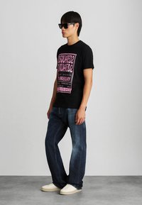 Modelo masculino con gafas de sol negras, camiseta gráfica negra con texto rosa, jeans sueltos azules y zapatillas blancas, de pie de lado.