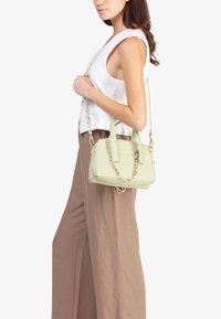Borsa a mano verde chiaro con una superficie liscia, manici corti e un accento a catena. La borsa presenta una forma strutturata e una tracolla regolabile.