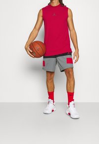 Rode mouwloze shirt met een logo, grijze shorts met rode accenten, en witte sneakers met rode details, terwijl hij een basketbal vasthoudt.