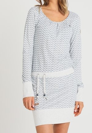 Nainen, jolla on harmaa- ja valkoisella siksak-kuviolla varustettu pitkähihainen mekko, jossa on kiristysnauha vyötäröllä ja käännetyt hihansuut.