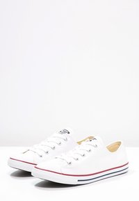 Baskets en toile blanches avec fermeture à lacets, semelles en caoutchouc et un accent rayé rouge et marine le long du bas. Design plat, sans motifs visibles.