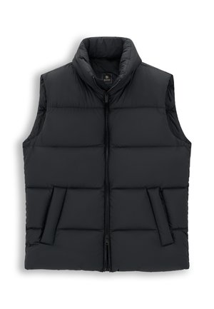 Un homme porte un gilet noir matelassé sur un pull beige et une chemise blanche, avec un pantalon crème et des chaussures marron, sur un fond uni.
