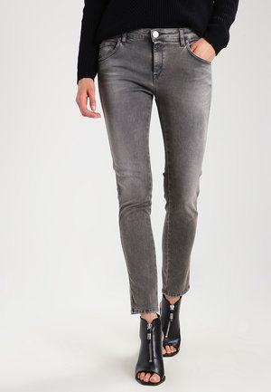 Person trägt ausgewaschene graue Slim-Fit-Jeans, schwarze offene Knöchelstiefel mit Frontreißverschlüssen und einen schwarzen Pullover, Hand in der Tasche.