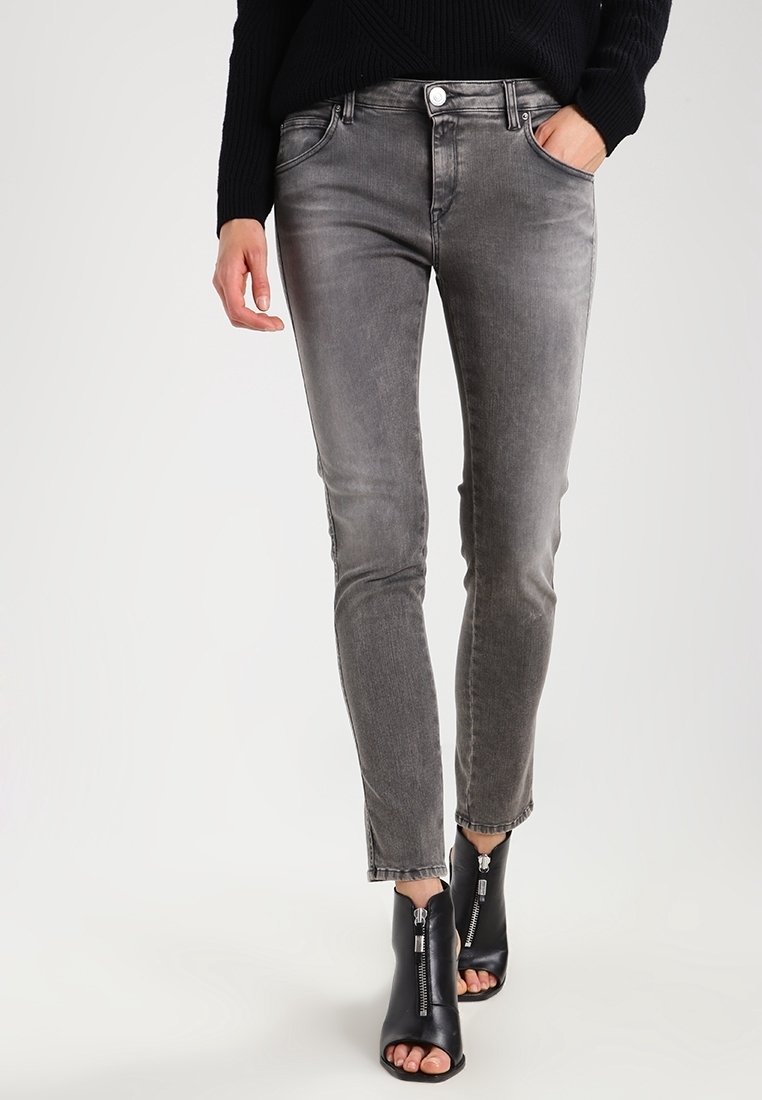 Personne portant un jean slim gris délavé, des bottines noires à bouts ouverts avec des fermetures éclair à l'avant, et un pull noir, main dans la poche.
