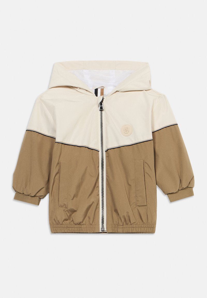 Giacca leggera con cappuccio in crema e marrone. Presenta una zip frontale, polsini elastici e due tasche laterali. Include una toppa con il logo.