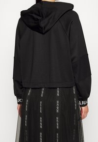 Czarna krótka bluza z kapturem, z długimi rękawami i prążkowanym wykończeniem na dole. Mankiety ozdobione są napisem "LIU JO SPORT" w kontrastowym kolorze.