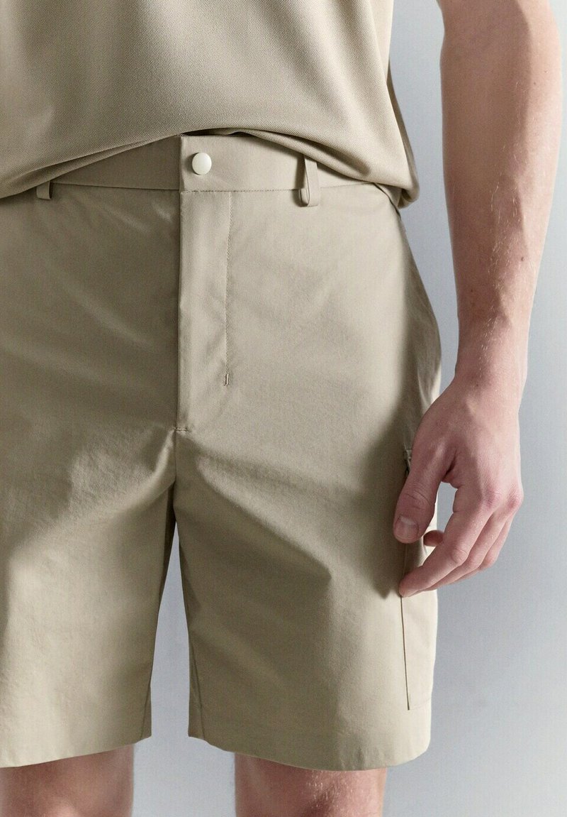 Shorts beige ajustés avec poches latérales portés par une personne avec une chemise beige rentrée, visibles du milieu de la cuisse à la taille.