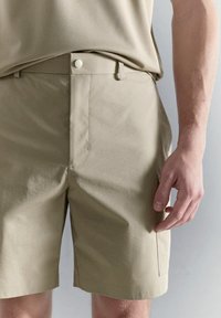 Pantalones cortos beige a medida con bolsillos laterales, llevados por una persona con una camisa beige metida por dentro, mostrados desde la mitad del muslo hasta la cintura.