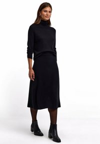 Schwarzer Strickrollkragenpullover kombiniert mit einem schwarzen knielangen Rock. Das Model trägt schwarze Knöchelstiefeletten, die einen eleganten, monochromen Look unterstreichen.