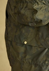 Kapiuotė tamsiai alyvuogių spalvos audinio su gėlių raštu. Yra matomas snapelis ir glotni tekstūra.