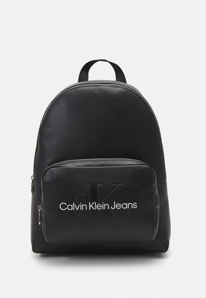 Zaino in pelle nera con una forma arrotondata, dotato di una tasca anteriore con zip e l'etichetta "Calvin Klein Jeans" e di un manico superiore per un facile trasporto.
