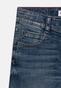 Niebieskie dżinsy mają klasyczny pięciokieszeniowy fason, widoczne przeszycia i miedziane nity. Tkanina ma zniszczoną fakturę z subtelnym wyblaknięciem.