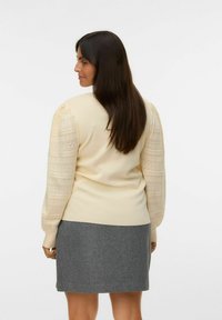Cremefarbener Pullover mit strukturierten Puffärmeln, kombiniert mit einem grauen Rock. Der Pullover hat einen glatten Strickkörper und komplizierte Ärmelmuster.