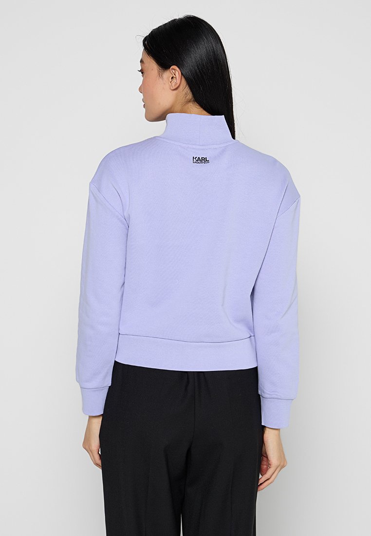 Sudadera corta de lavanda con cuello alto, que presenta puños y dobladillo ribeteados, y un logo negro en la parte posterior. Tejido suave y con textura.