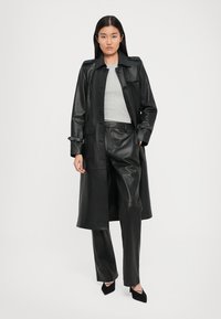 Black leather trench coat with button closure and adjustable belt, paired with black leather pants and gray top. Pointed black heels.