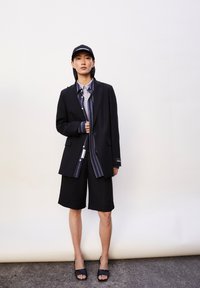 Schwarze Blazer mit strukturierten Schultern, kombiniert mit einem Hemd mit blauen und grauen Streifen, schwarzen Shorts und schwarzen Sandalen. Schwarze Kappe.
